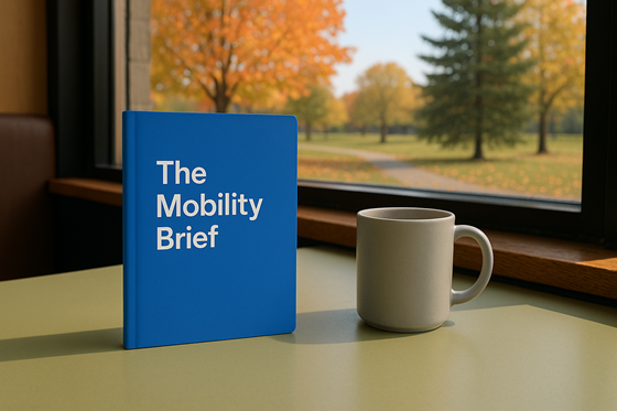 A photograph of a closed, blank notebook, laying flat on a table, next to a plain ceramic mug.  On the notebook is the title 'The Mobility Brief' with no other words, images, engravings or symbolism on the cover. The table is in a relaxed cafe environment, beside a window. Outside of the window there is a quiet park with a Sugar Maple and a White Pine. The weather outside is crisp and clear, with the first hints of red and gold appearing in the trees and a scattering of leaves on the ground.