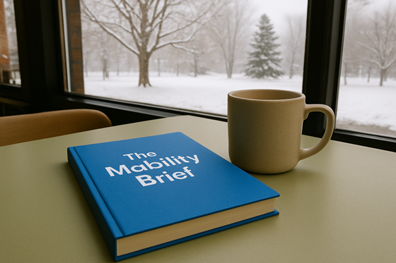 A photograph of a closed, blank notebook, laying flat on a table, next to a plain ceramic mug.  On the notebook is the title 'The Mobility Brief' with no other words, images, engravings or symbolism on the cover. The table is in a relaxed cafe environment, beside a window. Outside of the window there is a quiet park with a Sugar Maple and a White Pine. The weather outside is cold and grey, with a layer of snow covering the ground and clinging to the bare tree branches.