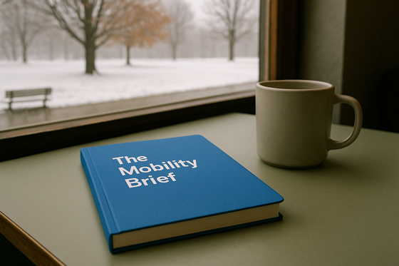 A photograph of a closed, blank notebook, laying flat on a table, next to a plain ceramic mug.  On the notebook is the title 'The Mobility Brief' with no other words, images, engravings or symbolism on the cover. The table is in a relaxed cafe environment, beside a window. Outside of the window there is a quiet park with a sugar maple and a white oak. The weather outside is cold and overcast, with a layer of snow blanketing the ground and clinging to the bare trees.