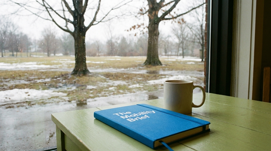 A photograph of a closed, blank notebook, laying flat on a table, next to a plain ceramic mug.  On the notebook is the title 'The Mobility Brief' with no other words, images, engravings or symbolism on the cover. The table is in a relaxed cafe environment, beside a window. Outside of the window there is a quiet park with a Sugar Maple and a White Oak. The weather outside is overcast and damp, with bare trees rising from the soggy, thawing ground where patches of old snow still cling.