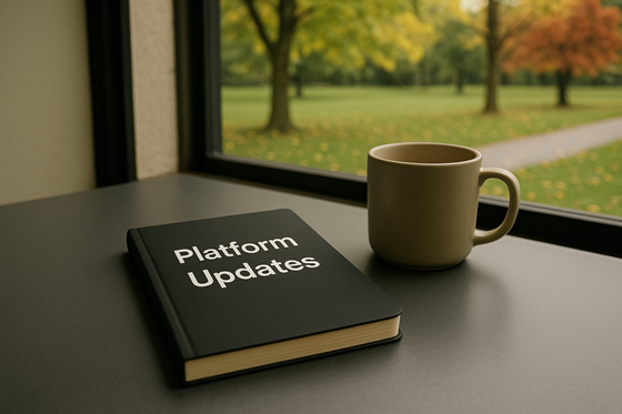 A photograph of a closed, blank notebook, laying flat on a table, next to a plain ceramic mug.  On the notebook is the title 'Platform Updates' with no other words, images, engravings or symbolism on the cover. The table is in a relaxed cafe environment, beside a window. Outside of the window there is a quiet park with a Sugar Maple and a Red Oak. The weather outside is clear and mild, with the leaves on the trees just beginning to turn yellow and scatter over the still-green ground.